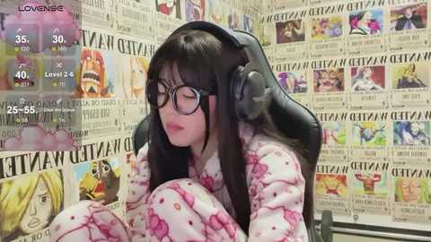 Media: Video of a young woman with long black hair, wearing glasses and a pink floral pajama set, seated in a gaming chair with large headphones. The background is filled with newspaper clippings featuring colorful anime characters.