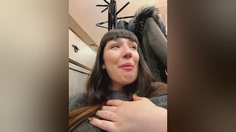 Media: A video of a Caucasian woman with shoulder-length dark hair, wearing a gray sweater, sitting on a black leather couch with a brown jacket draped over her. The background features a beige wall and a black ceiling light fixture.