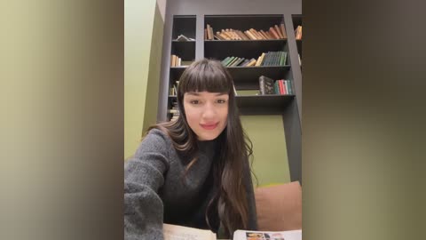 Media: Video of a young woman with long dark hair and bangs, wearing a dark gray sweater, sitting in a library with a bookshelf filled with books behind her.