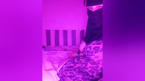 Media: Video of a person with dark hair, wearing a black mask, seated on a leopard-print bed, bathed in purple light. Background features vertical striped walls.