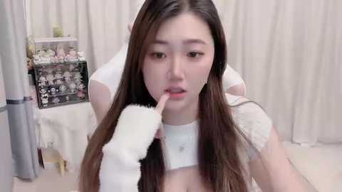 Media: Video of a young Asian woman with long, straight brown hair, wearing a white top, gently feeding a small white cat with a dropper. The background features a white curtain and a decorative shelf with small objects.