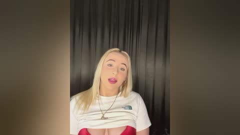 Media: Video of a blonde woman with fair skin, wearing a white t-shirt lifted to reveal a red bra, against a dark curtain background.