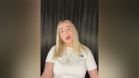 Media: Video of a blonde woman with fair skin, wearing a white T-shirt and pink lipstick, against a black curtain backdrop. She has a casual, slightly pouty expression.