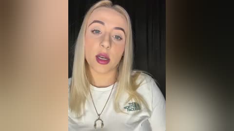 Media: Video of a young, blonde Caucasian woman with straight hair, wearing a white long-sleeve shirt, black necklace, and pink lipstick. She has a neutral expression. Background is dark.