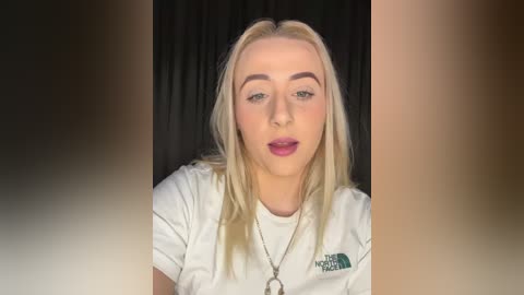 Media: Video of a young blonde woman with fair skin, wearing a white T-shirt, dark lipstick, and a silver necklace, against a black curtain background.