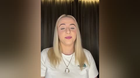 Media: Video of a young, fair-skinned woman with long, straight blonde hair, wearing a white t-shirt, pink lipstick, and a silver necklace, against a dark curtain background.