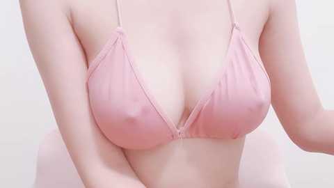 Media: Video of a light-skinned woman wearing a pink, halterneck bikini top, highlighting her ample cleavage and smooth, pale skin. Background is a plain, off-white wall.