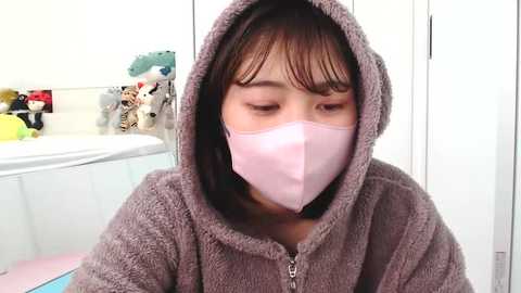 Media: Video of an Asian woman with short black hair wearing a light pink face mask and a brown, hooded robe, standing in a modern bathroom with white tiles and a white bathtub.