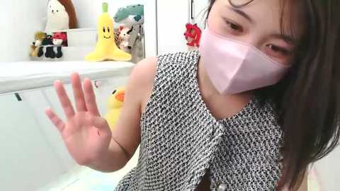 Media: Video of an East Asian woman with long black hair, wearing a pink surgical mask and a black and white houndstooth dress, making a hand gesture in a brightly lit room filled with stuffed toys and a yellow rubber duck.