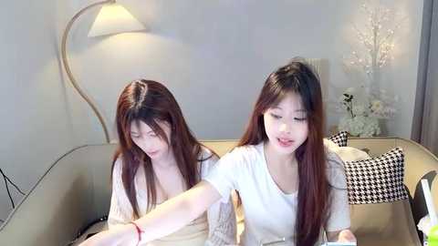 Media: Video of two Asian women with long brown hair, wearing white tops, sitting on a beige couch in a modern living room.