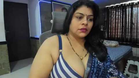 Media: Video of a plus-sized South Asian woman with dark hair, wearing a blue and white striped bra, sitting in a modern office.