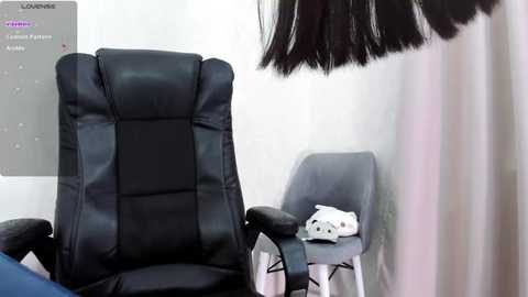 Media: Video of a sleek black leather office chair, a white plush toy, and a modern grey chair in a minimalist room with white walls and a black ceiling fan.