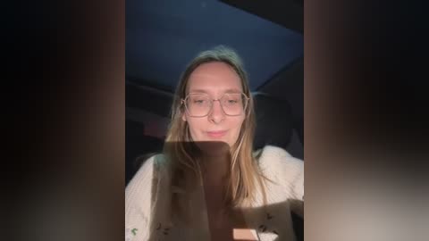 Media: Video of a fair-skinned woman with long, straight blonde hair, wearing glasses and a white cardigan, looking out of a car window in low light.