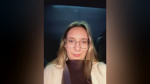 Media: Video of a young Caucasian woman with light brown hair, wearing glasses, a beige cardigan, and a black top, set against a dimly lit background.