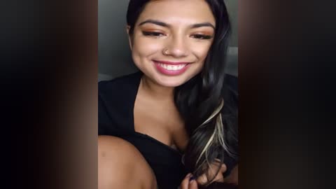 Media: Video of a smiling woman with medium skin tone, long black hair, and dark brown eyes, wearing a black top, holding a black feather in her hand.