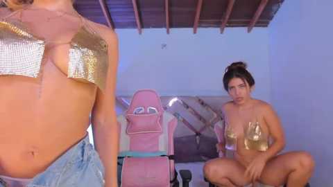 Media: Video of two women: One with a bikini top and denim shorts, the other topless with a pink gaming chair and a glass of wine in the background.