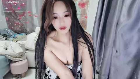 Media: Video of a young East Asian woman with long, straight black hair, fair skin, and large breasts, wearing a black and white polka dot top, sitting on a bed with scattered clothes and pink floral decor.