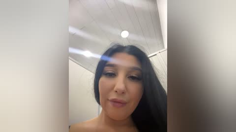 Media: A close-up video of a young woman with long black hair, fair skin, and full lips, making a pouty face. She is indoors with a white ceiling and fluorescent lights.