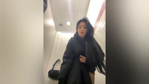 Media: Video of a woman in a public restroom, wearing a black coat and holding a black handbag, standing in a narrow, brightly lit hallway with white walls and a trash bin.