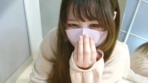 Media: Video of an Asian woman with long, straight brown hair and a pale pink surgical mask, covering her nose and mouth, sitting indoors. She wears a beige sweater.