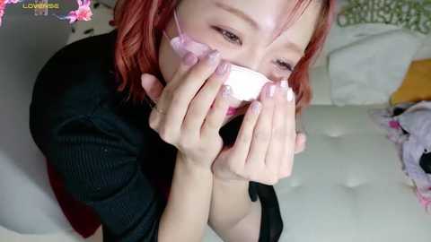 Media: Video of a young Asian woman with red hair, wearing a black sweater, covering her face with a white cloth. She sits on a white bed, surrounded by colorful pillows.