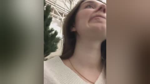 Media: Video of a smiling woman with straight brown hair and fair skin, wearing a white knitted sweater, standing in a greenhouse with greenery and metal structures in the background.