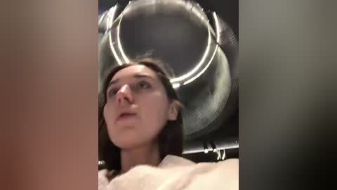 Media: Video of a woman with light skin and brown hair, wearing a beige top, looking upwards at a large, round, wire-framed mirror. The background is dimly lit, with a blurred, dark, possibly metallic surface.