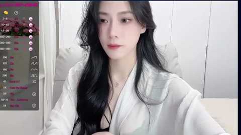 Media: Video of a young East Asian woman with long black hair, fair skin, and a slender physique, wearing a white robe, sitting indoors.