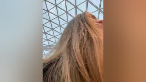 Media: Video of a young woman with long, straight, blonde hair, looking up, against a modern geometric glass ceiling with grid pattern.