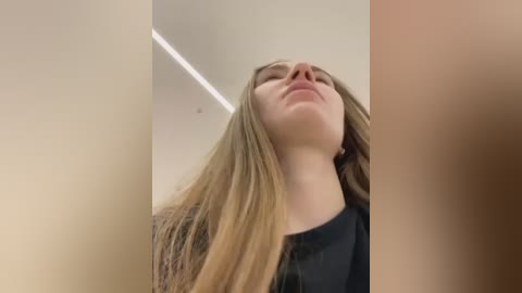 Media: Video of a young woman with long, straight blonde hair, wearing a black top, gazing upwards with closed eyes, taken indoors with minimalistic decor and a ceiling light.