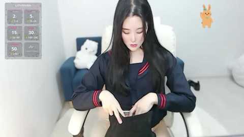 Media: Video of an East Asian woman with long black hair, wearing a navy school uniform, sitting on a white chair. She's holding a black purse. Background includes a plush toy, a temperature gauge, and a cartoon character.