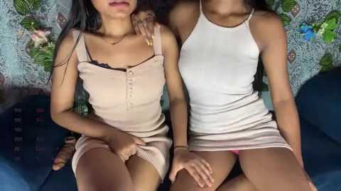 Media: Video of two young women with light brown skin, wearing tight, revealing beige and white tank dresses, sitting closely on a dark blue couch, with a floral-patterned wall background.