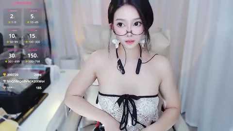 Media: Video of an East Asian woman with fair skin, wearing glasses, a white lace dress, and black tassel earrings, seated indoors.