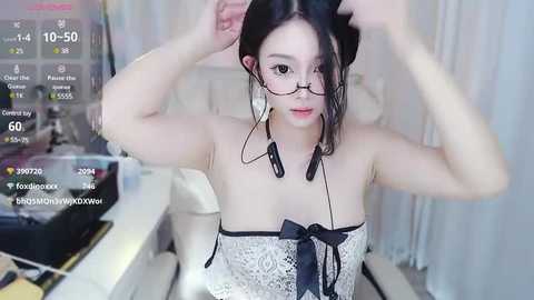 Media: A video of a young Asian woman with fair skin, wearing glasses, a lace top, and headphones, adjusting her hair, in a modern, brightly lit room with a white background.