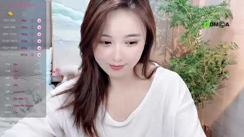 Media: Video of an East Asian woman with long brown hair, wearing a white off-shoulder top, smiling softly. Background includes a green plant and a blurred ocean-themed painting.