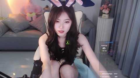 Media: Video of a young Asian woman in bunny ears, black lingerie, and a chair, with a gray sofa and flowers in the background.