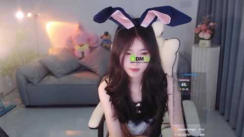 Media: Video of a young East Asian woman with long dark hair, wearing a black top and bunny ears, sitting in a modern living room. Background includes a grey couch, plush toys, and a digital thermometer.