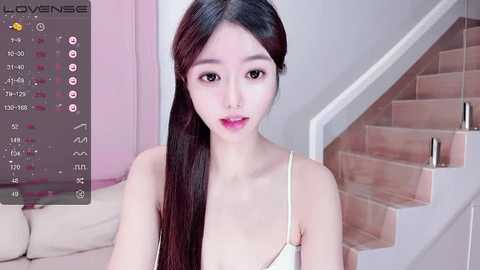 Media: Video of an Asian woman with long black hair, wearing a white spaghetti-strap top, standing on a beige sofa in a minimalist room with pink accents.