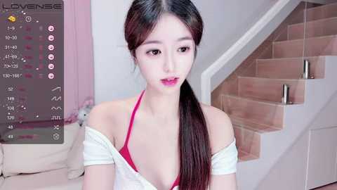 Media: Video of an East Asian woman with fair skin, long black hair in a ponytail, wearing a pink bra and off-white shirt, standing in a modern living room with stairs and plush toys in the background.