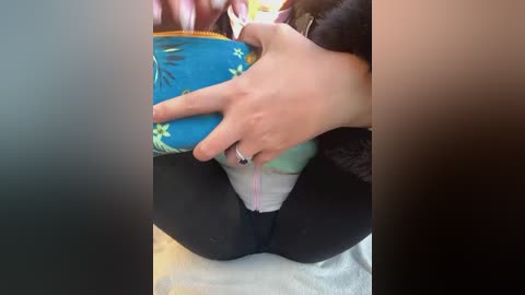 Media: A video shows a person sitting, holding a blue and green backpack with yellow flower patterns. They wear black leggings and a white top, revealing a glimpse of their underwear.