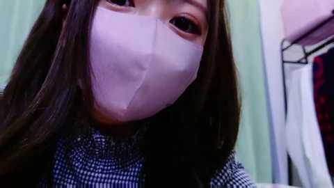 Media: Video of an Asian woman with long, dark hair, wearing a pink surgical mask, a black and white checkered shirt, and standing in a room with a hanging red plaid shirt and pale green wall.