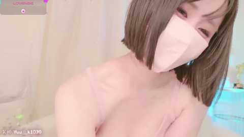 Media: Video of a young East Asian woman with a bob haircut and light skin, wearing a face mask and a pink bra, indoors with blurred background.