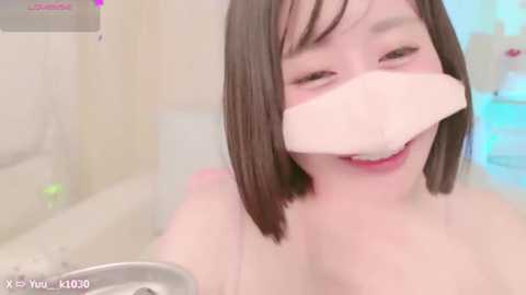 Media: A video of a smiling, light-skinned Asian woman with straight, shoulder-length brown hair, wearing a white bandage across her nose, sitting in a bathtub, with blurred bathroom background.