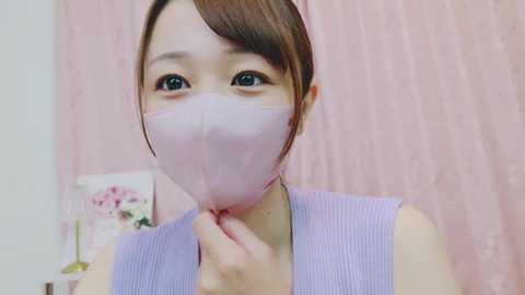 Media: A video of an Asian woman with fair skin and brown hair, wearing a light purple sleeveless top and a light pink surgical mask, standing against a pink, textured curtain background.