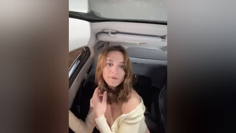 Media: Video of a young woman with shoulder-length light brown hair, wearing a white off-shoulder top, sitting in the backseat of a car, looking directly at the camera.