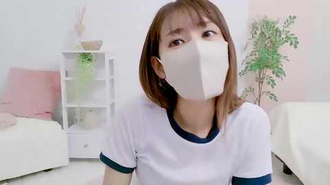 Media: Video of an Asian woman with shoulder-length brown hair, wearing a white T-shirt with navy blue trim, and a white face mask, sitting in a minimalist, pastel-colored bedroom with a white bed, a plant, and a shelf.