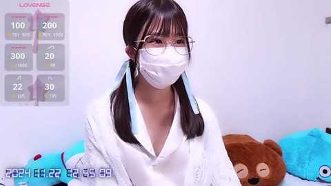 Media: Video of an East Asian woman with straight black hair, wearing glasses, a white surgical mask, and a white robe, seated against a plain white background with a teddy bear.