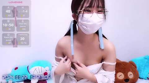 Media: A video of an East Asian woman with long black hair, wearing a white off-shoulder dress, face mask, and glasses, holding her chest, set against a light blue background with a plush toy.