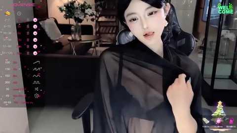 Media: Video of an Asian woman with fair skin, long black hair, and delicate features, wearing a sheer black dress, sitting in a modern living room.