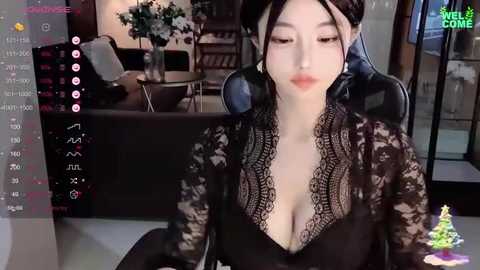 Media: Video of a young Asian woman with pale skin, dark hair, wearing a black lace top, sitting on a black gaming chair in a modern living room with a TV and a Christmas tree.